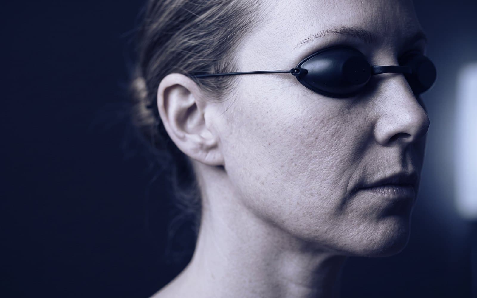 A woman's cheek and jaw in profile wearing protective goggles, light raking across the skin