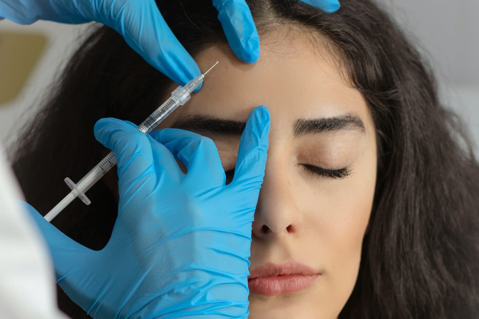 A provider in blue gloves placing a neuromodulator injection at a patient's brow, her eyes closed